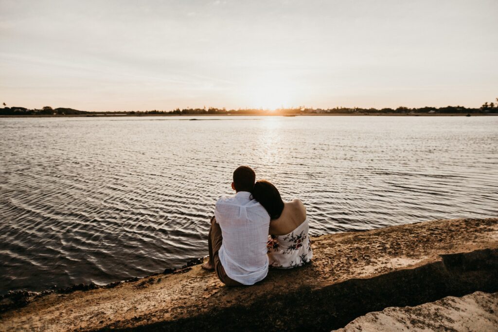 Back view unrecognizable female resting head on boyfriends shoulder while sitting together on stony river coast during romantic sunset-Relationship After a Loss