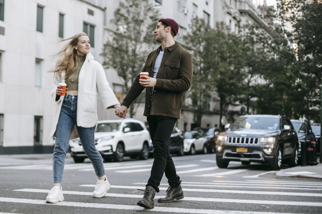 Young couple wearing warm jackets and with paper cups of hot drinks crossing road holding hands and looking at each other-Keep the Romance Alive
