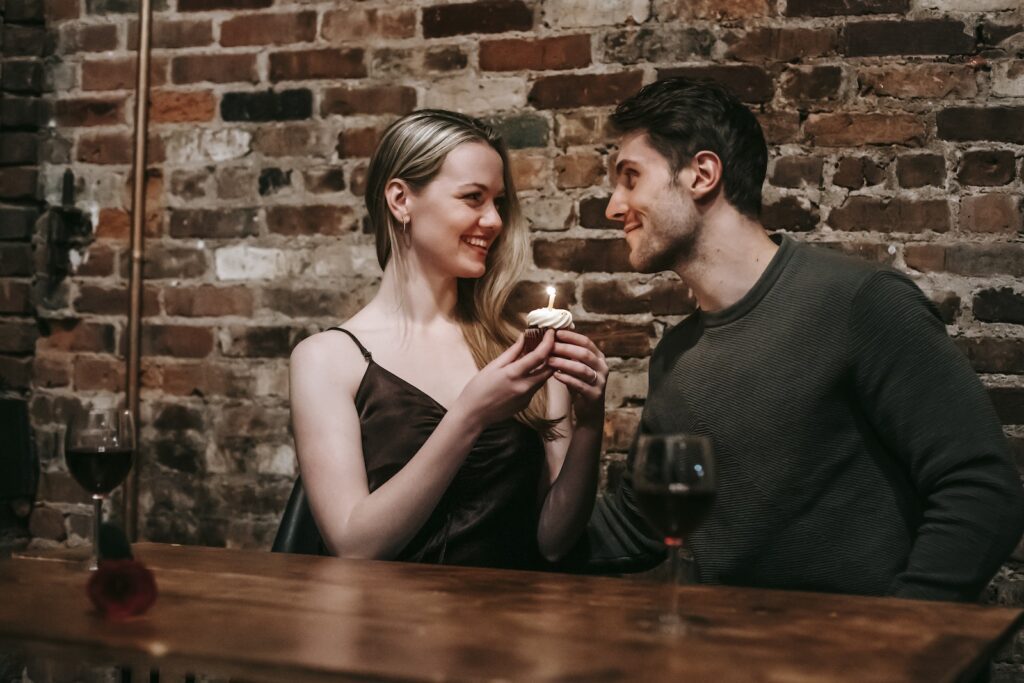 Side view of loving couple sitting near brick wall at wooden table with cupcake with burning candle looking at each other while celebrating anniversary in cafe-You're Ready for a Serious Relationship