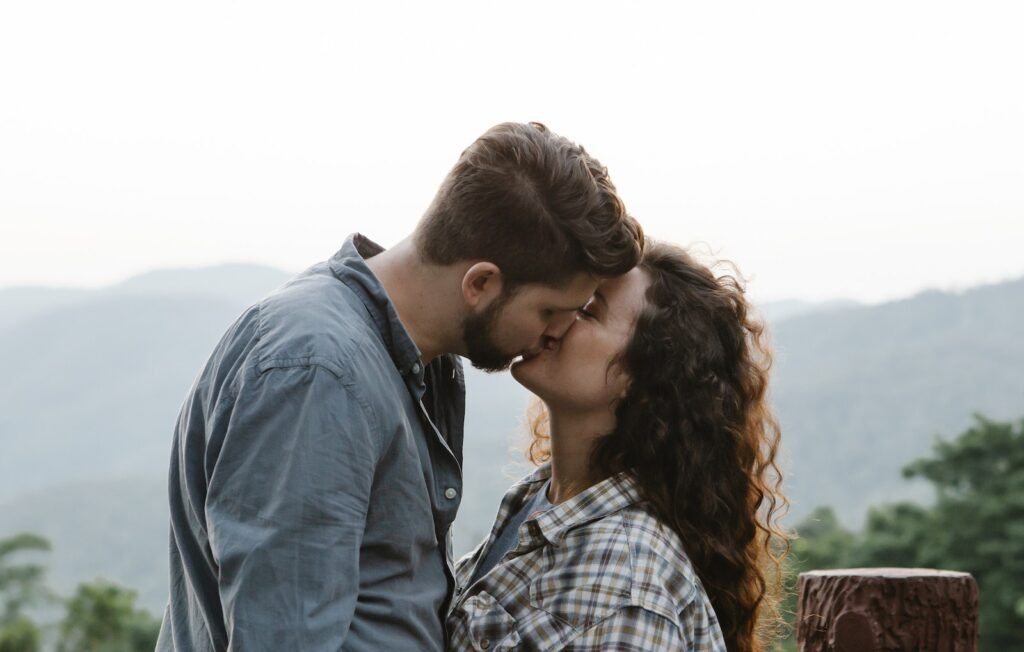 Side view of young loving couple in casual outfits kissing each other during romantic date in mountainous countryside-Flirting 