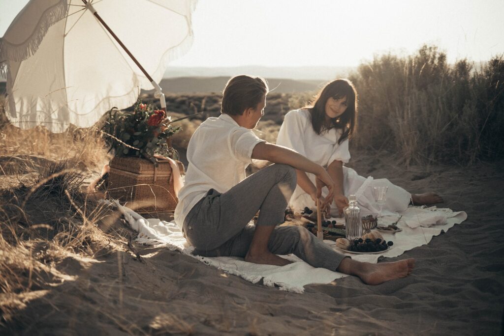 Young loving couple having romantic picnic sitting on white blanket with food and drinks under white umbrella-Long-Term Committed Relationship