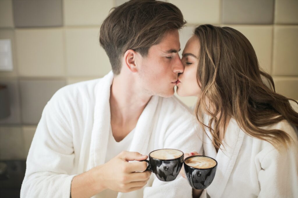 Photo of Couple in White Bathrobes Kissing While Holding Black Ceramic Cups of Coffee Couples Who Love Sports