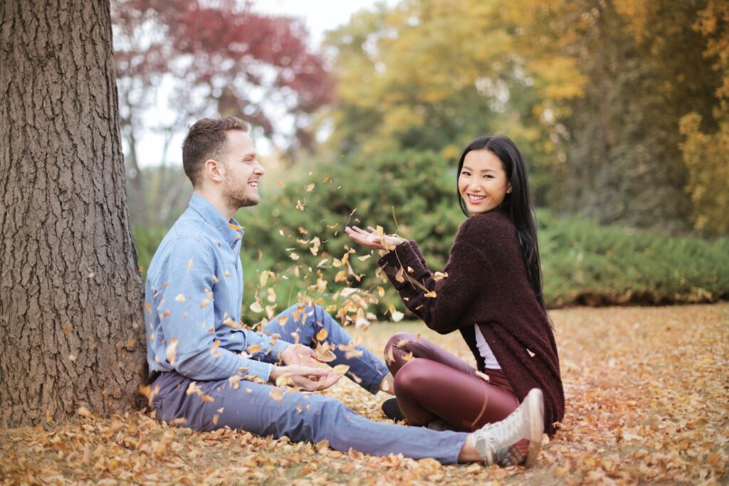 Side view of cheerful couple in casual clothes sitting in park and having fun while scattering dry leaves on autumn day-guy's attention