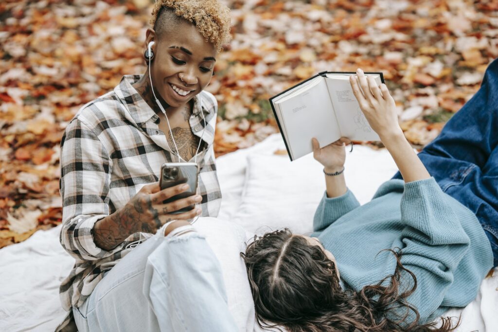 Anonymous female reading book while lying on plaid near African American girlfriend browsing smartphone and listening music in autumn nature on blurred background Love Overload