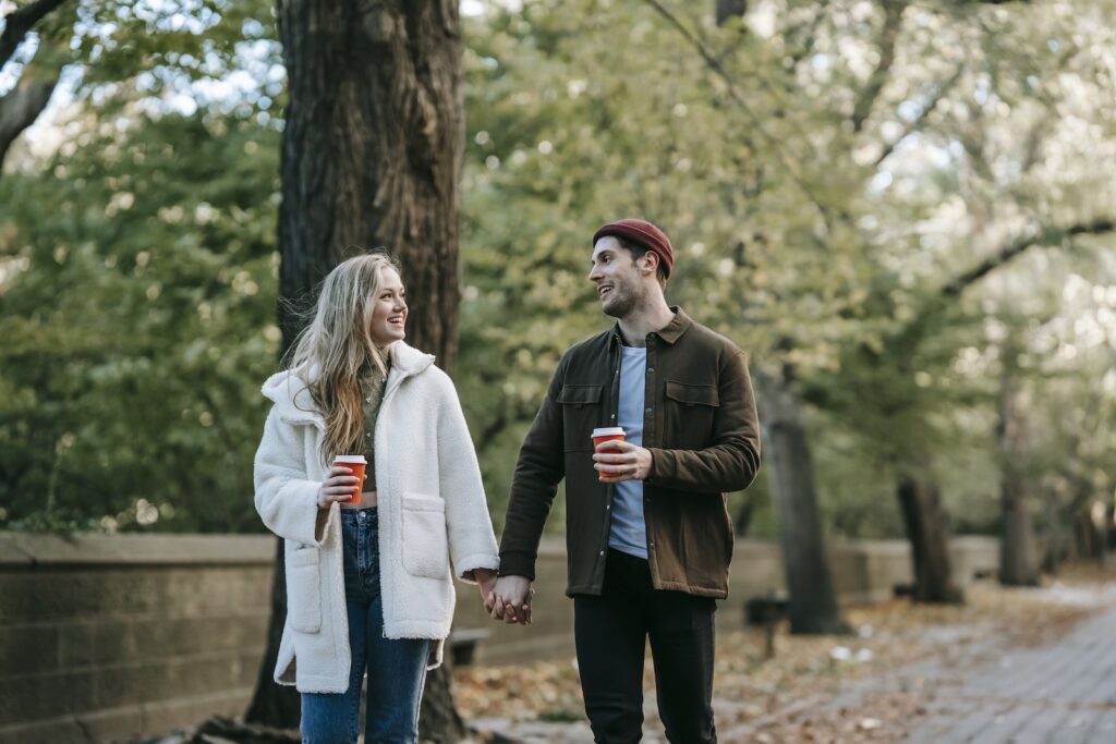 Happy stylish young man and woman in warm clothes holding hands and chatting while walking in autumn park and drinking takeaway coffee Heartbreak