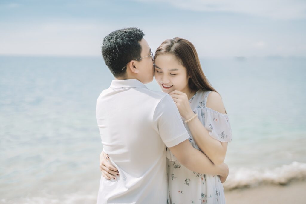 Ethnic couple in elegant outfits standing on sandy coast near ocean and embracing each other gently while man kissing woman in cheek in sunny summer day under blue sky Committed Relationship