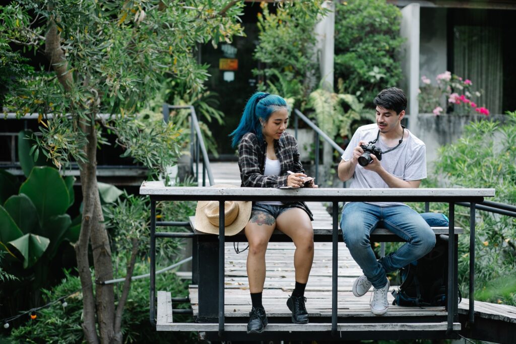 Young male photograph in casual clothes adjusting camera and talking with girlfriend model with blue hair and in checked jacket sitting on wooden bench in suspension footbridge in green garden You're Ready for a Committed Relationship