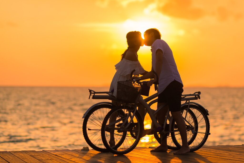 Photography of Man Wearing White T-shirt Kissing a Woman While Holding Bicycle on River Dock during Sunset-Get a Guy to Ask Me Out
