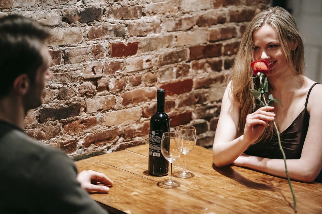 Young couple in elegant clothes sitting at wooden table near brick wall in light restaurant while enjoying date with red rose and wine near glasses and looking at each other-Falling for You