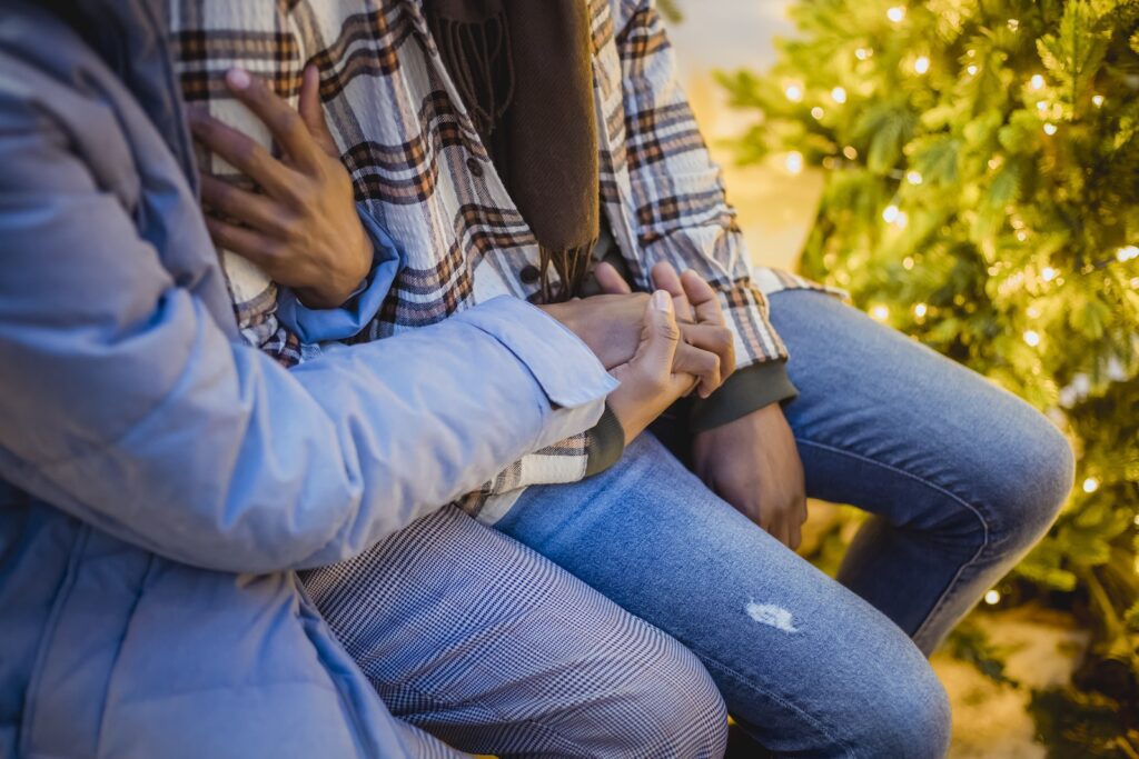 Unrecognizable romantic couple in holding hands and cuddling while sitting on street near Christmas tree with glowing lights during romantic date-Lasting 
