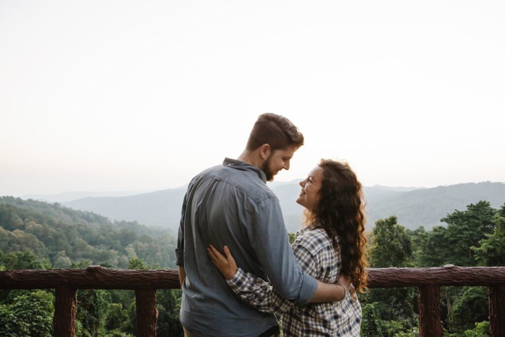 Back view of happy young romantic couple cuddling and looking at each other while standing on terrace against picturesque lush forest in mountains-Transformation