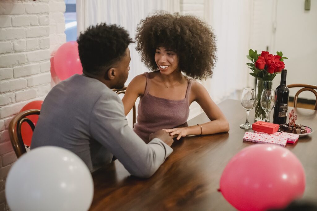 Young black couple in elegant clothes talking while sitting at table with wine bottle and glasses near flowers bouquet in vase and balloons in home while looking at each other-Effective Communication Strategies