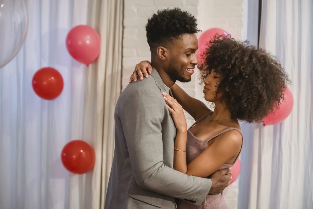Side view of young black couple in elegant clothes standing in bright room and cuddling while dancing together with closed eyes near pink balloons on wall and curtains-Confidence in Dating