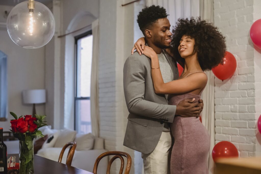Smiling young African American couple in elegant outfits embracing and dancing while standing in bright apartment near balloons on wall and table with bouquet of roses in vase-Resolve Conflicts