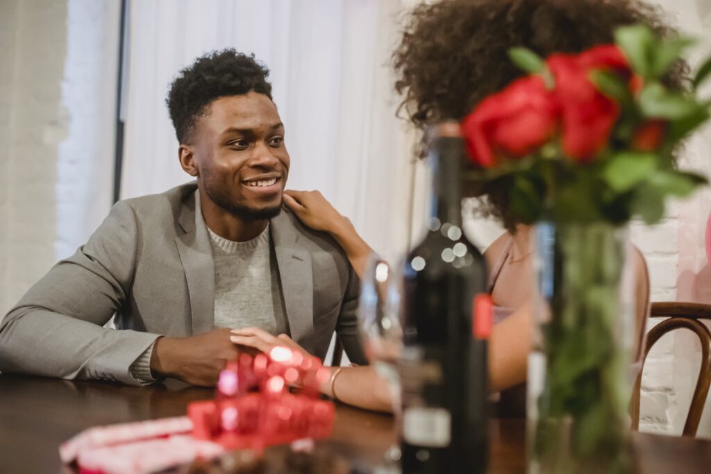 Happy young black couple in elegant clothes talking while holding hands at table with wine bottle and glasses near flowers bouquet in vase at home while looking at each other-Supporting Your Spouse